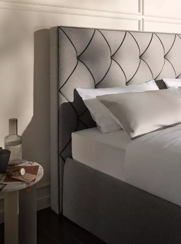 Elegantes graues Bett mit diamantförmigem Polsterkopfteil, weißer Bettwäsche, beigen Kissen